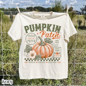 Pode incluir: Uma t-shirt de manga curta em tom off-white com uma estampa gráfica de uma abóbora laranja, folhas verdes e o texto PUMPKIN Patch, FRESH FROM THE PATCH, FARM FRESH, LOCALLY GROWN, AUTUMN PICKED, PUMPKINS, CORN MAZE e APPLE CIDER.
