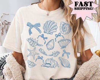 May include: A white t-shirt with a blue seashell and starfish design. The shirt has a crew neck and short sleeves. The design includes various seashells, starfish, and bows.