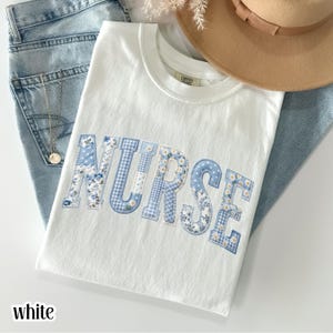 Könnte beinhalten: Weißes T-Shirt mit dem Wort "NURSE" in blauen Patchwork-Buchstaben. Die Buchstaben weisen Blumen-, Vichy- und Gänseblümchenmuster auf. Das T-Shirt ist auf einer blauen Jeans gefaltet, mit einer goldenen Halskette und einem beigen Hut im Hintergrund.