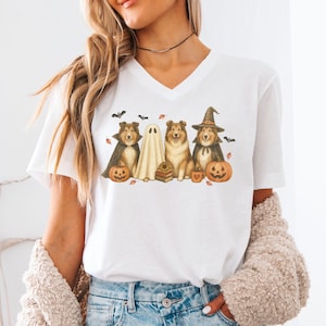May include: White V-neck t-shirt featuring a Halloween-themed graphic. The design includes illustrations of dogs dressed as a witch, a ghost, and a dog in a cape, along with pumpkins and bats. The shirt is made of a soft material.