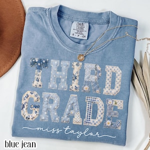 May include: A blue jean Comfort Colors t-shirt with the words "THIRD GRADE" in patchwork letters. The letters are decorated with floral and patterned fabrics. The name "miss taylor" is written below the words. A gold necklace is draped across the shirt.