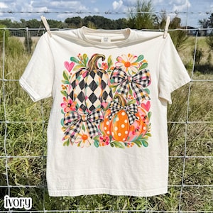 Könnte beinhalten: Ein elfenbeinfarbenes Kurzarm-T-Shirt mit einem Grafikdruck von zwei Kürbissen, einer mit schwarz-weißem Rautenmuster und einer orangefarbenen mit weißen Punkten, akzentuiert durch schwarz-weiß karierte Schleifen, rosa Herzen und bunte Blumen.