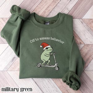 Op de afbeelding: Militairgroen sweatshirt met de tekst "Off to assess behavior". De afbeelding toont een kikker met een kerstmuts op een scooter. Het sweatshirt is effen van kleur.