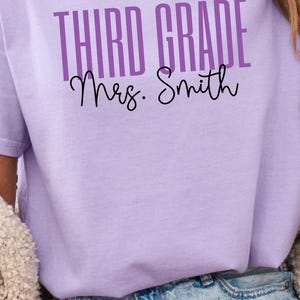 May include: A collection of short-sleeved crew neck t-shirts in blue, light purple, and off-white. Each shirt features the text "THIRD GRADE" in a bold sans-serif font and "Mrs. Johnson" or "Mrs. Smith" in a black cursive script font.