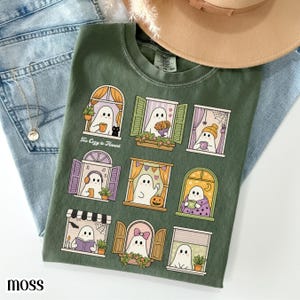 Könnte beinhalten: Ein olivgrünes T-Shirt mit einem Grafikdruck von neun Fenstern, in denen jeweils ein weißer Geist verschiedene Aktivitäten ausübt. Der Text "Too Cozy to Haunt" ist in Weiß nahe dem oberen linken Fenster gedruckt. Das T-Shirt hat einen Rundhalsausschnitt.