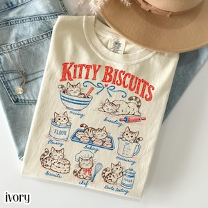 以下が含まれることがあります： アイボリー色のTシャツで、猫がパン作りをしている赤と青のグラフィックが描かれています。KITTY BISCUITSという文字と、mixing、kneading、flouring、baking、measuring、biscuits、chef、taste testingというラベルがあります。クルーネックでComfort Colorsのラベルが付いています。