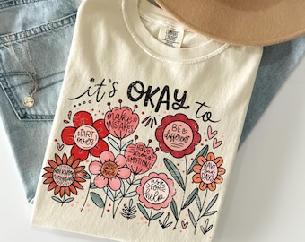 Op de afbeelding: Ivoor T-shirt met een bloemenontwerp en de tekst "it's OKAY to." De bloemen bevatten berichten als "make mistakes" en "be different." Het shirt wordt getoond met een blauwe jeans en een beige hoed.
