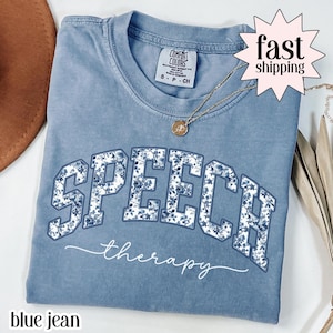 以下が含まれることがあります： 大きな花柄の文字で「SPEECH」と、その下に筆記体で「therapy」と書かれた、ブルーデニムカラーのTシャツ。ペンダント付きのゴールドネックレスが見えます。シャツはComfort Colorsブランドです。