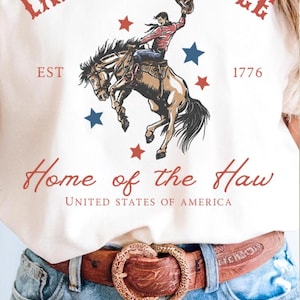 以下が含まれることがあります： カウボーイのグラフィック、赤い文字で「Land of the Yee」と「Home of the Haw」、そして年号「1776」がプリントされた明るい色のTシャツ。「4th of July Cowboy Shirt」の文字も含まれています。