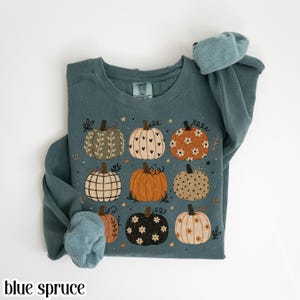 May include: A blue spruce crewneck sweatshirt featuring a graphic print of nine pumpkins in various patterns, including floral, polka dot, heart, and plaid designs, interspersed with small stars and hearts. The inner neck label reads Comfort Colors.