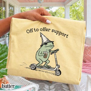 May include: A pale yellow t-shirt featuring a graphic of a green frog wearing a pointed hat, riding a white scooter, and holding a beverage. The text "Off to offer support" is printed in black above the frog illustration on the fabric.