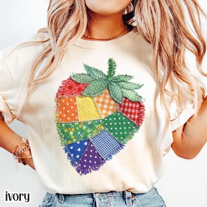Puede incluir: Sudadera de color marfil con un diseño de fresa de patchwork colorido. La fresa está compuesta por varios patrones de tela, incluyendo lunares, cuadros y estampados florales. La palabra "ivory" está impresa en la parte inferior de la imagen.