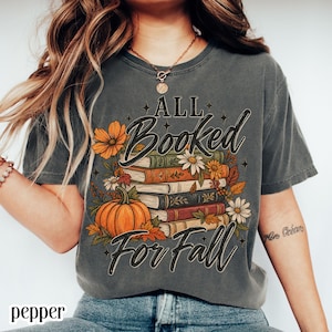 Puede incluir: Una camiseta de manga corta gris carbón con un estampado gráfico de una pila de libros, una calabaza y flores de otoño. El texto "ALL Booked For Fall" está impreso en negro. La camiseta tiene un aspecto vintage y desgastado.