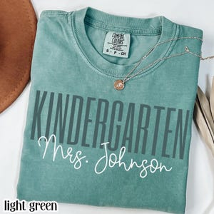 May include: Light green t-shirt with the words "KINDERGARTEN Mrs. Johnson" printed in gray and white. A gold necklace with a pendant is draped across the shirt. The shirt is folded and placed on a white surface.