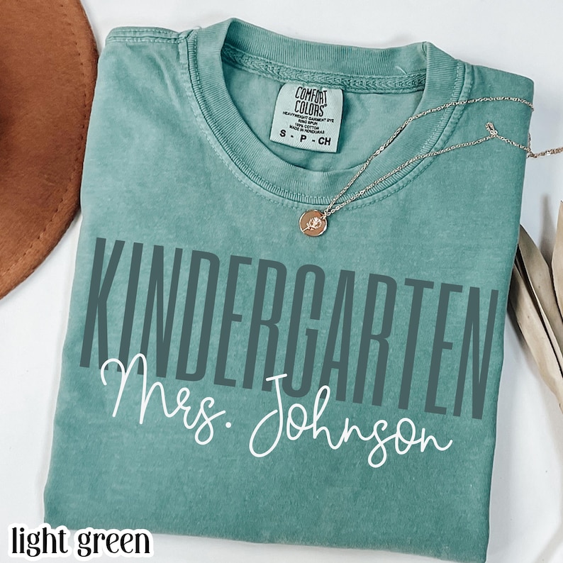 May include: Light green t-shirt with the words "KINDERGARTEN Mrs. Johnson" printed in gray and white. A gold necklace with a pendant is draped across the shirt. The shirt is folded and placed on a white surface.
