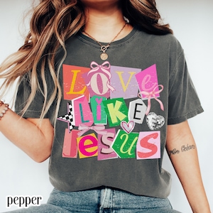 May include: Dark gray t-shirt with the words "LOVE LIKE JESUS" in a colorful, collage-style design. The design includes pink bows, a checkered star, and a disco heart. The shirt is a casual, comfortable style.