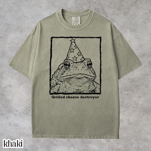 Puede incluir: Camiseta caqui con una ilustración en blanco y negro de un sapo con un gorro de fiesta. El texto "Grilled cheese destroyer" está debajo de la imagen. La camiseta tiene cuello redondo y mangas cortas.