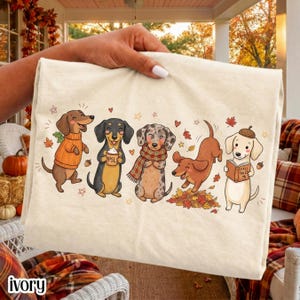 May include: A cream-colored t-shirt featuring a graphic illustration of five dachshunds in autumn-themed attire. The dogs are depicted wearing sweaters and scarves, holding a coffee cup labeled PSL, playing in leaves, and reading a book titled Fall Tales.
