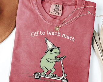Può includere: T-shirt Comfort Colors color cremisi con la scritta "Off to teach math". La maglietta presenta un ranocchio dei cartoni animati con un cappello da mago che guida un monopattino. Una collana d'argento con un ciondolo è drappeggiata sulla maglietta.