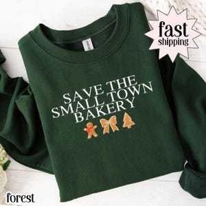 May include: Forest green sweatshirt with the text "SAVE THE SMALL TOWN BAKERY" in white. Below the text are embroidered images of a gingerbread man, a bow, and a Christmas tree. The words "fast shipping" are in a starburst.
