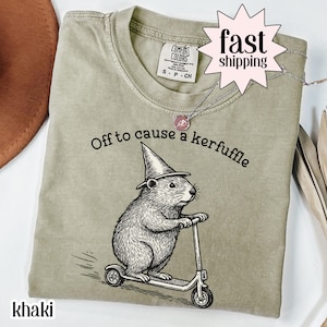 Könnte beinhalten: Khaki-farbenes T-Shirt mit dem Schriftzug "Off to cause a kerfuffle" und einer Grafik eines Nagetiers mit Zaubererhut auf einem Roller. Eine silberne Halskette mit rosa Anhänger ist ebenfalls zu sehen.