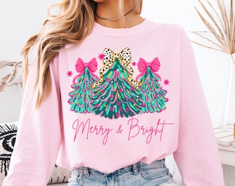 Puede incluir: Sudadera rosa claro con un diseño festivo de árbol de Navidad. El gráfico presenta tres árboles turquesa con lazos rosas y un lazo con estampado de leopardo. Las palabras "Merry & Bright" están escritas en rosa.
