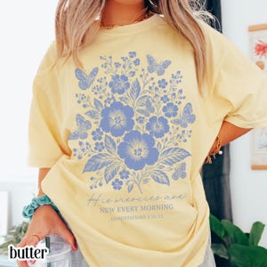 Könnte beinhalten: Ein buttergelbes Kurzarm-T-Shirt mit einer großen blauen Grafik aus Blumen und Schmetterlingen. Unter dem Blumenmotiv steht der Text: His mercies are NEW EVERY MORNING LAMENTATIONS 3:22-23 in einer Kombination aus Schreib- und Blockschrift.