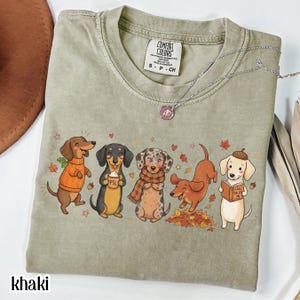 May include: A sage green t-shirt featuring a graphic of five dachshunds in autumn attire. The dogs are depicted wearing sweaters and scarves, holding a pumpkin spice latte, playing in leaves, and reading a book titled Fall Tales.
