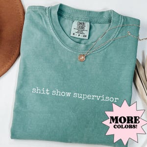 May include: A teal blue t-shirt with the text "shit show supervisor" printed in white. The shirt has a distressed look and is available in more colours.