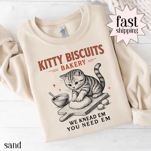 Puede incluir: Sudadera de color arena con un gráfico de un gatito amasando masa en una tabla de madera. El texto "KITTY BISCUITS BAKERY" está encima del gatito, con "WE KNEAD EM YOU NEED EM" debajo. La sudadera tiene cuello redondo.