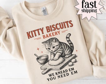 May include: Sand-colored sweatshirt featuring a graphic of a kitten kneading dough on a wooden board. The text "KITTY BISCUITS BAKERY" is above the kitten, with "WE KNEAD EM YOU NEED EM" below. The sweatshirt has a crew neck.
