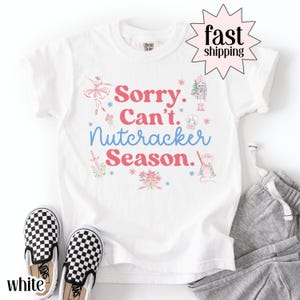 Puede incluir: Camiseta blanca con el texto "Sorry. Can't. Nutcracker Season." en rosa y azul. La camiseta presenta ilustraciones de una bailarina, un cascanueces y un ratón. Se ven zapatos sin cordones a cuadros blancos y negros.