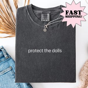Puede incluir: Una camiseta gris oscuro con el texto "protect the dolls" impreso en blanco en la parte delantera.