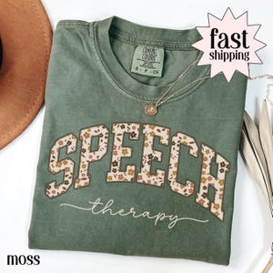 May include: A moss green t-shirt with the word "SPEECH" in a floral design and the word "therapy" in a cursive font. The shirt has a Comfort Colors tag and a small necklace. The text "fast shipping" is in a pink starburst.