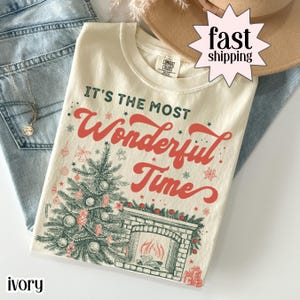 Ivory Christmas shirt with “It’s the Most Wonderful Time” text, featuring a decorated Christmas tree, fireplace, ornaments, and holiday accents in vintage-style artwork