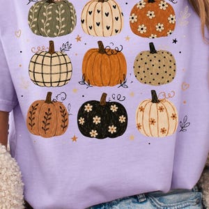 May include: A t-shirt featuring a graphic print of nine pumpkins in various colors and patterns, including floral, polka dot, plaid, and heart designs, accented with small stars and leaves. The shirt is shown in white, light purple, and moss green.
