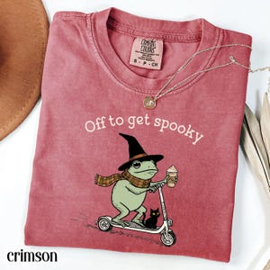 May include: A crimson short-sleeved t-shirt featuring a graphic of a green frog wearing a witch hat and plaid scarf, riding a scooter with a black cat and holding a coffee cup. The text reads Off to get spooky. The shirt has a Comfort Colors label.