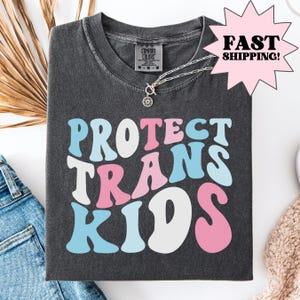Puede incluir: Camiseta gris oscuro con las palabras "PROTECT TRANS KIDS" en una fuente retro, con letras en azul claro, rosa y blanco. Un collar plateado con un colgante de sol está en la camisa. La camisa está doblada sobre una superficie blanca.