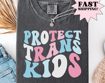 Peut inclure: T-shirt gris foncé avec l'inscription "PROTECT TRANS KIDS" dans une police rétro, avec des lettres bleu clair, roses et blanches. Un collier argenté avec un pendentif soleil est sur le t-shirt. Le t-shirt est plié sur une surface blanche.