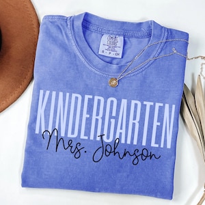 May include: A periwinkle Comfort Colors t-shirt with the words "KINDERGARTEN Mrs. Johnson" printed in white and black. A gold necklace with a pendant rests on the shirt. A brown hat is visible in the upper left corner.