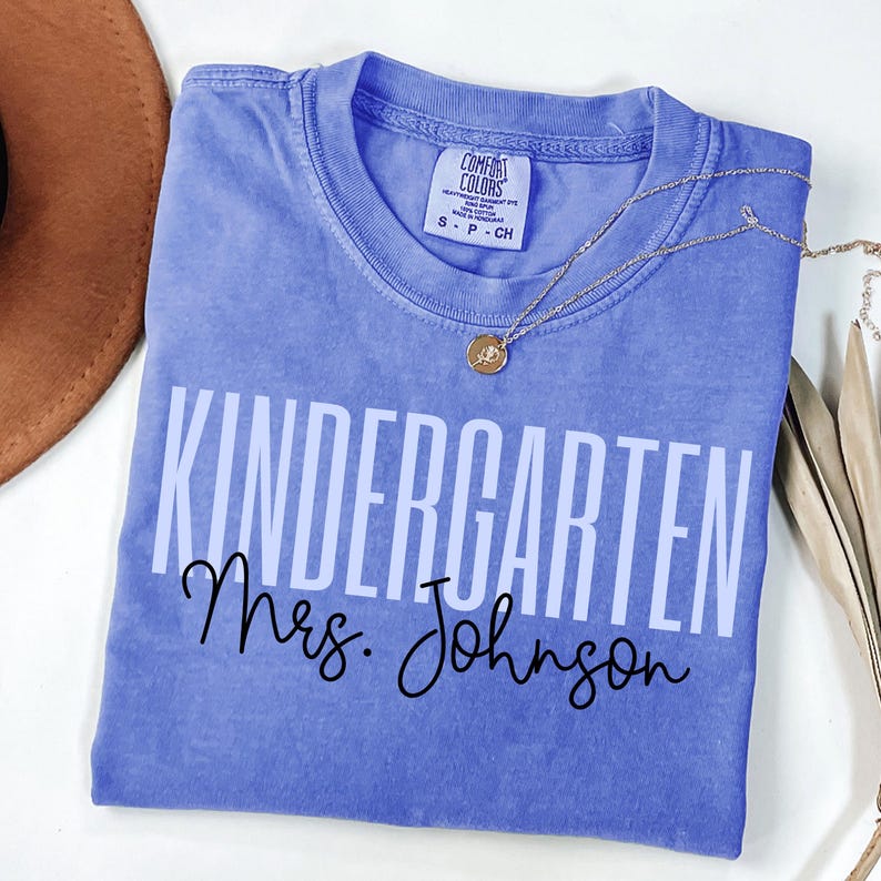 May include: A periwinkle Comfort Colors t-shirt with the words "KINDERGARTEN Mrs. Johnson" printed in white and black. A gold necklace with a pendant rests on the shirt. A brown hat is visible in the upper left corner.