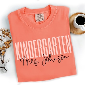 May include: Coral-colored t-shirt with the word "KINDERGARTEN" in large, light pink block letters and "Mrs. Johnson" in black cursive. The shirt is a Comfort Colors brand and is next to a cup of coffee.