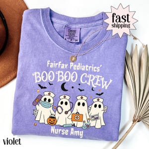 Könnte beinhalten: Ein violettes Comfort Colors T-Shirt mit dem Text "Fairfax Pediatrics' BOO BOO CREW" und Geistern als Krankenschwestern. Das Design umfasst Fledermäuse, einen Kürbis und "Nurse Amy". Eine goldene Halskette liegt auf dem T-Shirt.