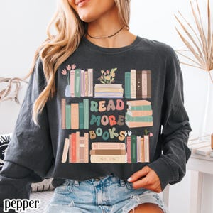 May include: Dark gray long-sleeve sweatshirt with a colorful graphic of stacked books and the text "READ MORE BOOKS". The design includes pastel-colored books and floral accents, creating a cozy, book-themed aesthetic.