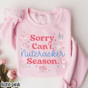 May include: Light pink sweatshirt with the text "Sorry. Can't. Nutcracker Season." in red and blue. The sweatshirt features illustrations of a ballerina, a nutcracker, and other holiday-themed graphics. The sleeves are folded.