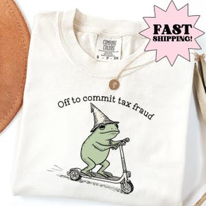Könnte beinhalten: Cremefarbenes T-Shirt mit dem Aufdruck "Off to commit tax fraud" und einem Cartoon-Frosch auf einem Roller, der einen Zaubererhut trägt. Eine goldene Halskette ist über dem T-Shirt drapiert.