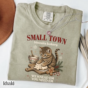 Könnte beinhalten: Ein khakifarbenes T-Shirt mit Rundhalsausschnitt und einer Grafik eines getigerten Kätzchens, das Teig auf einem Holzbrett knetet, neben einem Lebkuchenmann und einer Schüssel. Der Text lautet SMALL TOWN biscuits bakery und WE KNEAD EM YOU NEED EM.