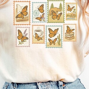 May include: A moss green, cream, and blue jean t-shirt with a graphic of orange monarch butterflies on postage stamps. The text "THE ONLY ORANGE MONARCHS I WANT" is above the stamps. The text "ANTI MONARCHY TESS" is below.