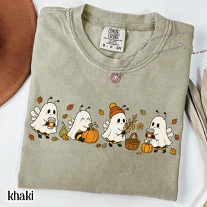 Puede incluir: Una camiseta de color caqui con un gráfico de cuatro fantasmas de dibujos animados vestidos de abejas, rodeados de hojas de otoño, calabazas, una cesta y tazas de café. La camiseta tiene cuello redondo y etiqueta Comfort Colors en el cuello.