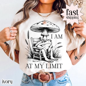 May include: Ivory-colored t-shirt featuring a black and white illustration of a frog sitting on a chair under a mushroom. The text "I AM AT MY LIMIT" is printed below the frog. The shirt has a crew neck.